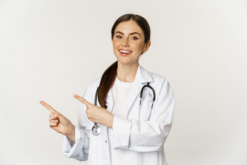Portrait of smiling young woman doctor, healthcare medical worker, pointing fingers left, showing clinic promo, logo or banner, standing over white background
